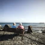 Back view full length faceless senior couple relaxing on bench on stone promenade and chatting while enjoying amazing peaceful seascape during sunny weather